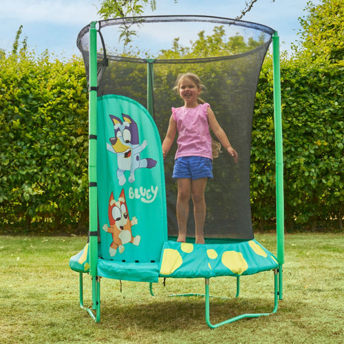 A young girl smiles and stands on a TP Toys Bluey UP 4.5ft Junior Trampoline with zip-free entry, a safety net, and Bluey graphics in a grassy backyard.