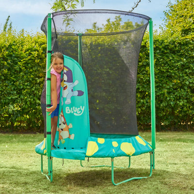 A young girl stands at the entrance of a TP Toys Bluey UP 4.5ft Junior Trampoline with a safety net and zip-free entry, set outdoors on a grassy lawn.