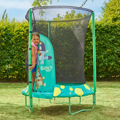 A young girl stands at the entrance of a TP Toys Bluey UP 4.5ft Junior Trampoline with a safety net and zip-free entry, set outdoors on a grassy lawn.