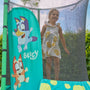 A young girl in a white floral dress stands on the TP Toys Bluey UP 4.5ft Junior Trampoline, which features Bluey and Bingo characters printed on its side panel.