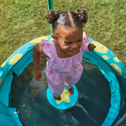 A young girl in a pink outfit with pigtails jumps on the TP Toys Bluey UP 4.5ft Junior Trampoline, featuring a blue and yellow design and zip-free entry, on a grassy lawn.