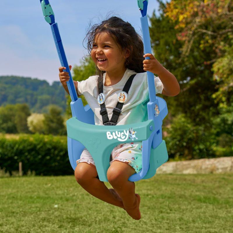 A young child smiles while swinging in a Bluey Quadpod Seat, part of the TP Toys Sherwood Wooden Double Swing with Roped Rapide Seat & Bluey Quadpod Seat, made from PEFC-certified wood, in an outdoor park setting.