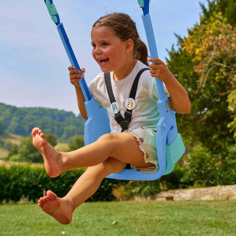 A brown-haired child in a white shirt smiles while swinging on the Bluey Quadpod Seat outdoors. The swing is part of the TP Toys Sherwood Wooden Double Swing with Roped Rapide Seat, made from PEFC certified wood.
