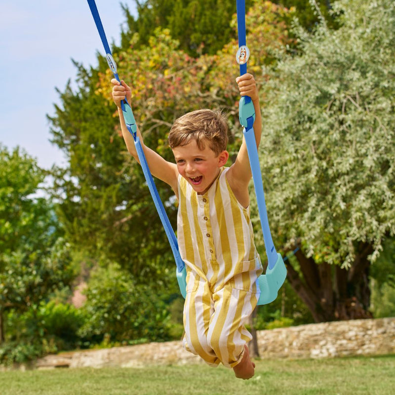 A young boy smiles while swinging on the Bluey Quadpod Seat of the TP Toys Sherwood Wooden Double Swing—featuring a PEFC certified roundwood frame, Roped Rapide Seat, and surrounded by trees and grass outdoors.