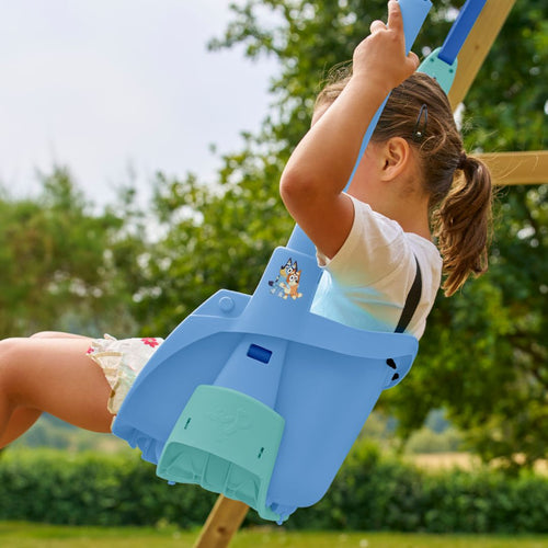 A young child sits in the TP Toys Bluey Quadpod® 4 in 1 Baby Swing Seat, holding the ropes on a wooden playset outdoors, featuring a Bluey cartoon sticker.