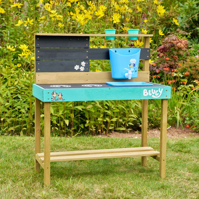 The TP Toys Bluey Wooden Mud Kitchen - FSC® certified features a wooden kitchen with Bluey logo, character stickers, blue tub, and plant pots, set in a garden with green grass and yellow flowers.