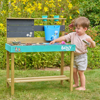 A young child enjoys the TP Toys Bluey Wooden Mud Kitchen—FSC® certified—playing in the garden on grass surrounded by yellow flowers.