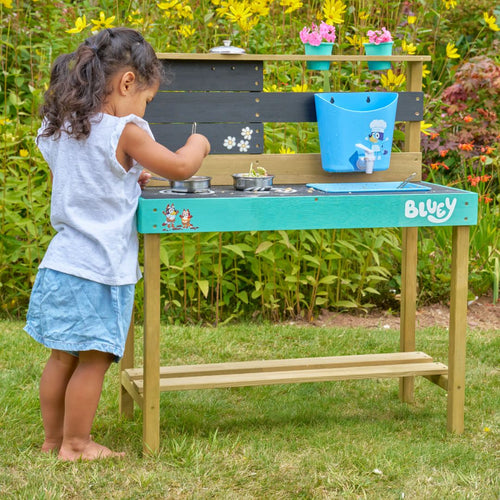 A young girl plays with the TP Toys Bluey Little Chef Wooden Mud Kitchen—FSC® certified—exploring pots, pans, and toy accessories in her imaginative outdoor setup, surrounded by green grass and flowering plants.