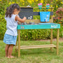 A young girl plays with the TP Toys Bluey Little Chef Wooden Mud Kitchen—FSC® certified—exploring pots, pans, and toy accessories in her imaginative outdoor setup, surrounded by green grass and flowering plants.