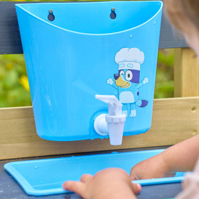 A child washes hands in a blue sink with tap, featuring a cartoon dog in a chef's hat—part of the TP Toys Bluey Wooden Mud Kitchen, FSC® certified.