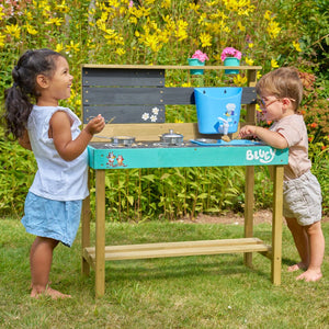 Two young children play at the TP Toys Bluey Little Chef Wooden Mud Kitchen - FSC® certified, featuring Bluey accents, surrounded by green grass and yellow flowers.