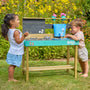 Two young children play at the TP Toys Bluey Little Chef Wooden Mud Kitchen - FSC® certified, featuring Bluey accents, surrounded by green grass and yellow flowers.