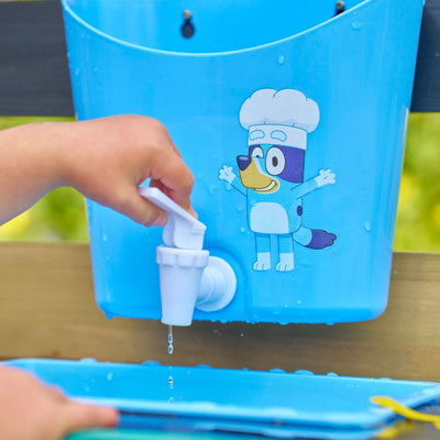 A child turns on a white tap attached to the TP Toys Bluey Wooden Mud Kitchen (FSC® certified), featuring a blue container with a cartoon dog in a chef hat; water drips from the tap.