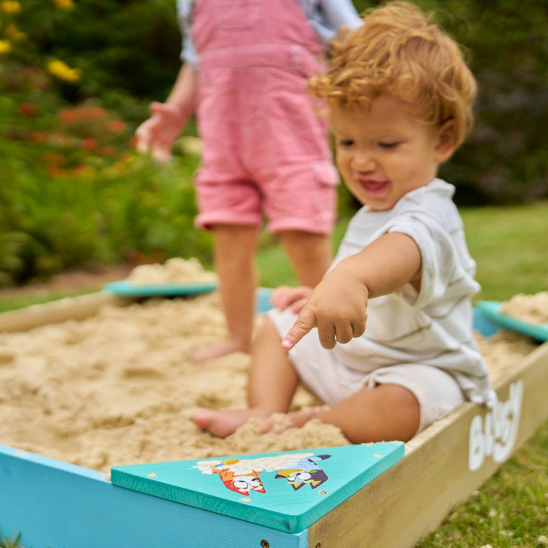 A young child sits in the TP Toys Bluey Wooden Sandpit with Cover, pointing at something in the sand, while another child in pink overalls stands nearby on the grass outdoors.
