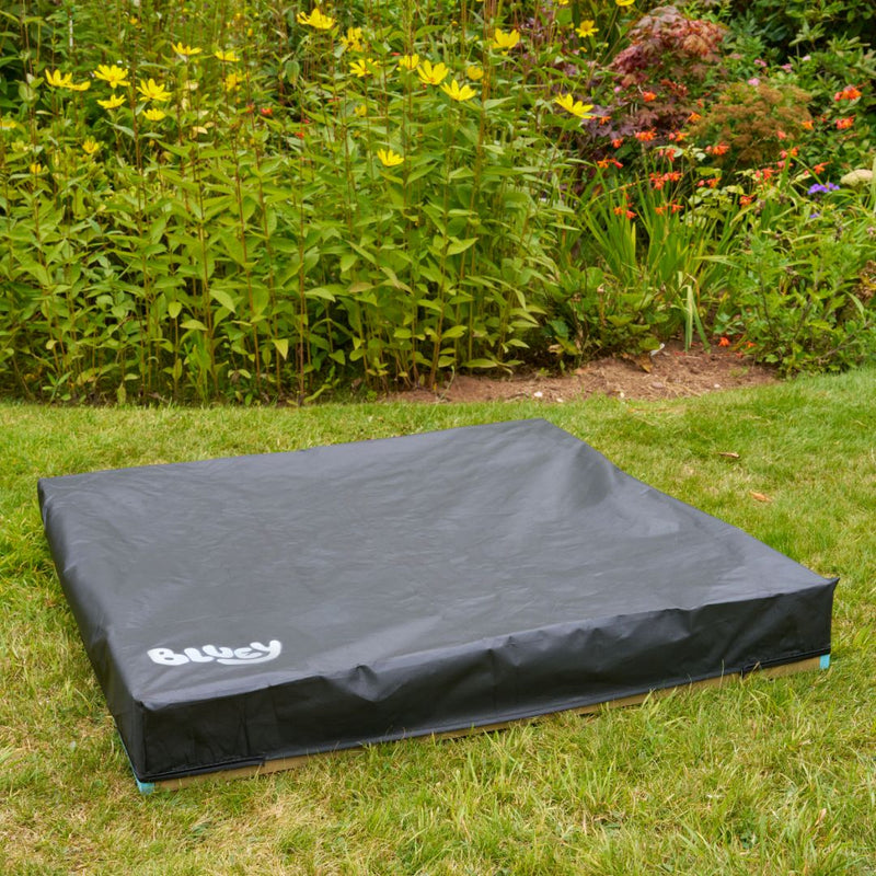 A black, square cover labeled "Olucy" sits atop the TP Toys Bluey Wooden Sandpit with Cover on grass, in front of a garden with yellow and green plants.