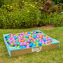 A TP Toys Bluey Wooden Sandpit with Cover sits on garden grass, filled with colorful plastic balls and surrounded by flowers and greenery.