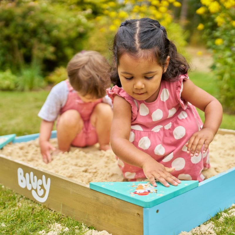 Two young children in pink outfits play outdoors with the TP Toys Bluey Wooden Sandpit with Cover. One lifts a blue corner lid while the other digs in the sand, both enjoying their sandbox fun under the sun.