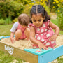 Two young children in pink outfits play outdoors with the TP Toys Bluey Wooden Sandpit with Cover. One lifts a blue corner lid while the other digs in the sand, both enjoying their sandbox fun under the sun.