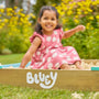A young girl in a pink polka dot dress smiles while sitting in the TP Toys Bluey Wooden Sandpit with Cover, surrounded by greenery and yellow flowers.
