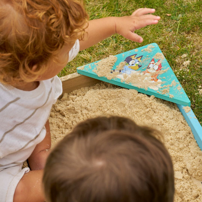 Two young children play in the TP Toys Bluey Wooden Sandpit with Cover, with sand on a cartoon-decorated corner and grass visible in the background.