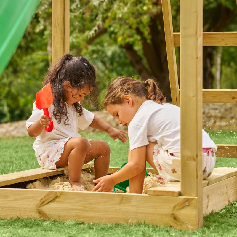 Two children play outdoors on the TP Toys Skywood Wooden Lookout Tower with 3m super wavy slide (FSC® certified), enjoying sand play with plastic shovels and buckets next to the wooden playset on a sunny day.