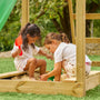 Two children play outdoors on the TP Toys Skywood Wooden Lookout Tower with 3m super wavy slide (FSC® certified), enjoying sand play with plastic shovels and buckets next to the wooden playset on a sunny day.