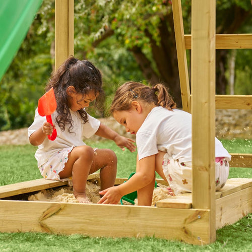 Two young children play in a wooden sandbox with plastic shovels and buckets, next to the TP Toys Skywood Wooden Lookout Tower with Ripple Slide & Double Swing Arm (FSC® certified), on grass surrounded by trees outdoors.