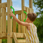 A young child in a yellow and white striped outfit holds green handles while climbing the TP Toys Skywood Wooden Lookout Tower with 3m Super Wavy Slide—an FSC® certified outdoor playset.