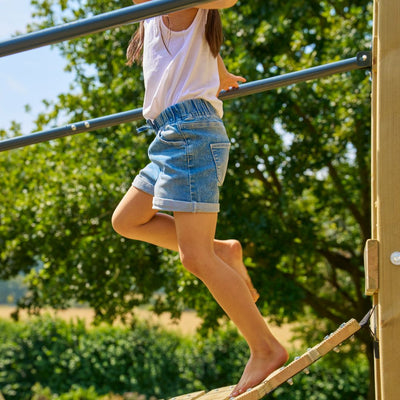A child in a white shirt and denim shorts balances barefoot on the TP Toys Skywood Wooden Tower Climbing Frame with slide, bridge, extra tower & double swing arm—FSC® certified—outdoors among trees and greenery.