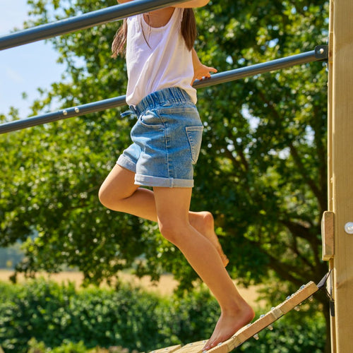 A child in a white shirt and denim shorts balances barefoot on the TP Toys Skywood Wooden Tower Climbing Frame with slide, bridge, extra tower & double swing arm—FSC® certified—outdoors among trees and greenery.