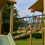 A young child walks across the TP Skywood Wooden SkyWalk Bridge by TP Toys, an FSC® certified modular climbing frame, with a green slide and grassy area in the background.
