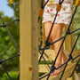 A child in floral shorts climbs the TP Toys Skywood Wooden SkyWalk Bridge (FSC® certified) barefoot, navigating the outdoor climbing net and wooden structure.