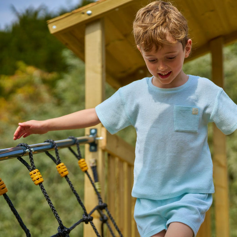 A young boy in a light blue t-shirt and shorts walks across the TP Toys Skywood Wooden SkyWalk Bridge—FSC® certified—in an outdoor playground.