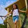 A young girl in a white shirt and denim shorts walks on the TP Skywood Wooden SkyWalk Bridge by TP Toys, part of a modular climbing frame at an outdoor playground. The bridge is FSC® certified.