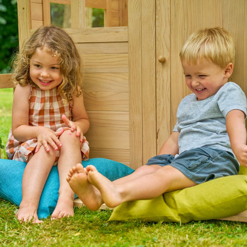 Two young children sit on colorful cushions outside the TP Toys Buttercup Cottage Wooden Playhouse with Floor, Verandah and Mud Kitchen, made from FSC® certified wood, smiling and laughing on a grassy lawn.