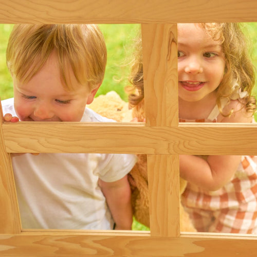 Two young children, one in a white shirt and one in a checkered dress, peek through the TP Toys Buttercup Cottage Wooden Playhouse with Floor, Verandah and Mud Kitchen—FSC® certified—with grass visible in the background.