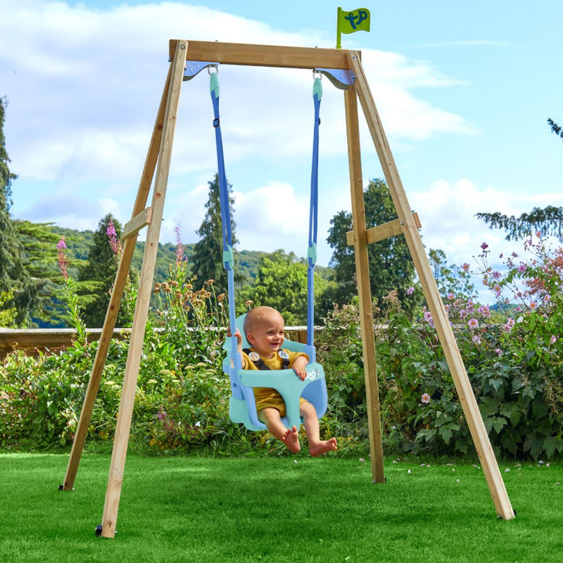A baby smiles in the TP Toys TP Forest Wooden Single Swing with Bluey Quadpod - FSC® certified, set up on grass amid trees and flowers.