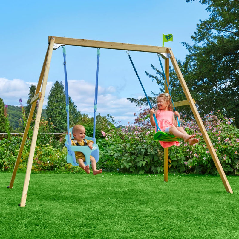 A young child and a baby enjoy the TP Toys TP Forest Wooden Double Swing Bluey with Quadpod and Rapide Seats (FSC® certified) in a grassy yard with flowers and trees in the background.