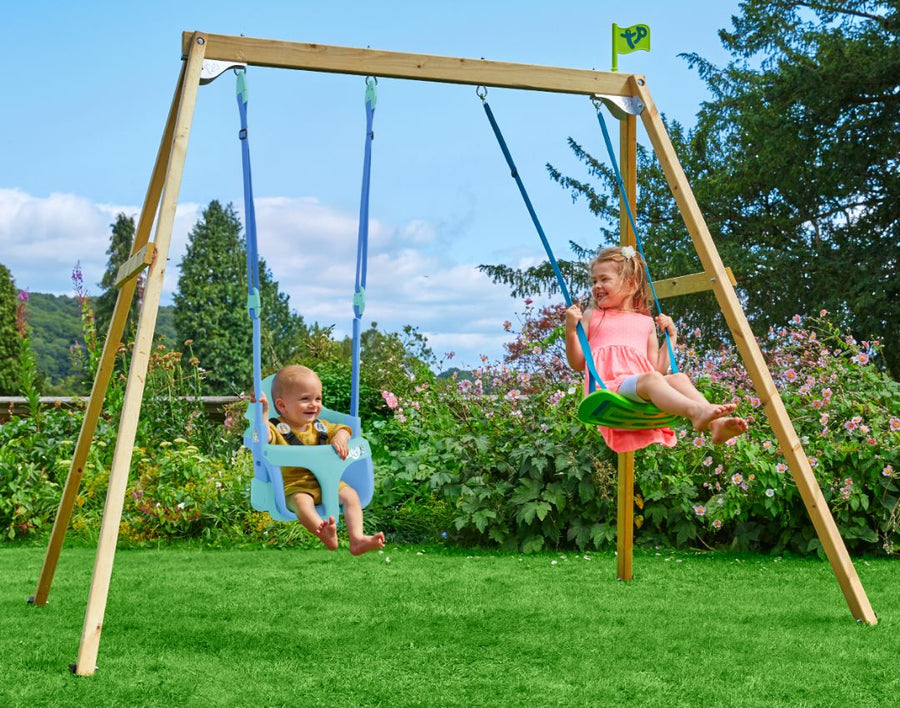 A young child and a baby enjoy the TP Toys TP Forest Wooden Double Swing Bluey with Quadpod and Rapide Seats (FSC® certified) in a grassy yard with flowers and trees in the background.