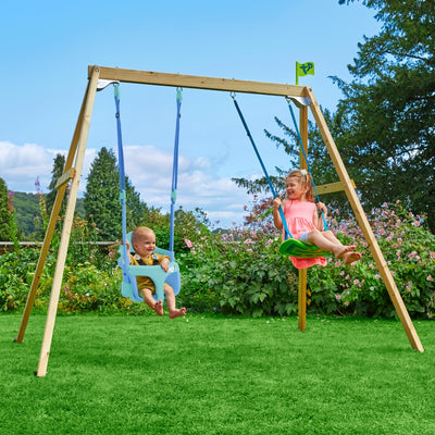 A baby and a young girl are sitting on swings attached to the TP Toys TP Forest Wooden Double Swing Bluey with Quadpod and Rapide Seats – FSC® certified, in a grassy garden with flowers and trees in the background.