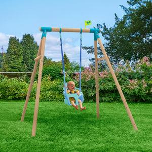 A baby sits in the TP Toys TP Sherwood Single Wooden Swing Set with Bluey Quadpod—PEFC certified—on a grassy lawn, surrounded by trees and bushes.