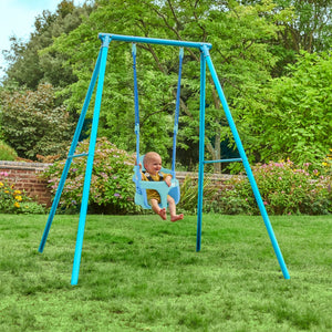 A baby sits in a blue TP Toys Bluey Quadpod on the TP Deluxe Single Metal Swing Set, suspended from its metal frame in a grassy garden with trees and flowers in the background.