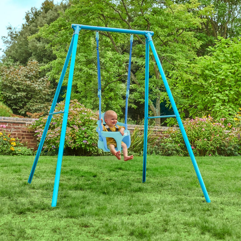 A baby sits in a blue TP Toys Bluey Quadpod on the TP Deluxe Single Metal Swing Set, suspended from its metal frame in a grassy garden with trees and flowers in the background.