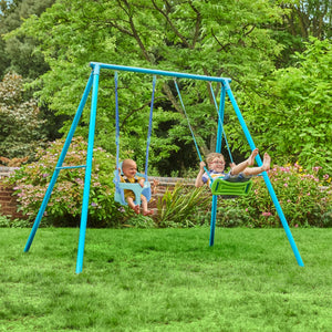 Two children swing on the TP Toys TP Deluxe Double Metal Swing with Rapide & Bluey Quadpod seats in a grassy backyard surrounded by trees and plants.