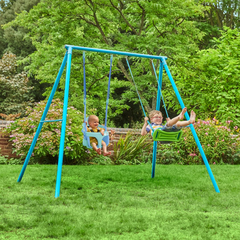 Two children swing on the TP Toys TP Deluxe Double Metal Swing with Rapide & Bluey Quadpod seats in a grassy backyard surrounded by trees and plants.