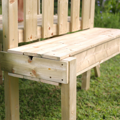 A close-up of a wooden outdoor bench on green grass in a garden, next to the TP Toys TP Forest Toddler Wooden Bluey Quadpod Swing Set & Slide (FSC® certified).