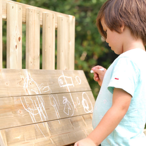 A child stands on the TP Forest Toddler Wooden Bluey Quadpod Swing Set & Slide by TP Toys, drawing with white chalk on the wooden outdoor surface, making simple shapes and figures.