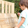 A child stands on the TP Forest Toddler Wooden Bluey Quadpod Swing Set & Slide by TP Toys, drawing with white chalk on the wooden outdoor surface, making simple shapes and figures.