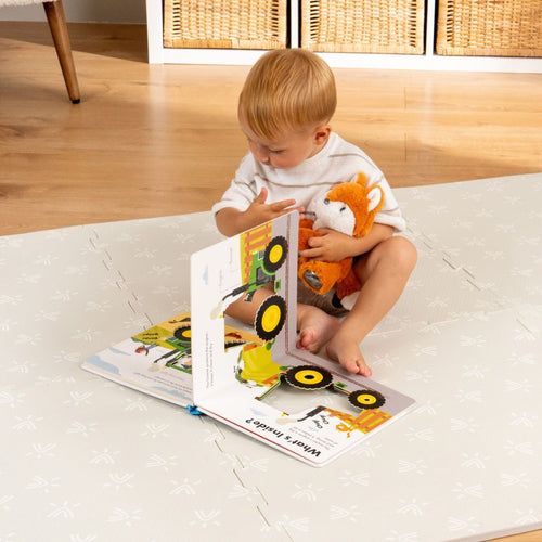 A young child sits on a TP Toys TP Active-Tots Safety Play Mat in a toddler play area, holding a stuffed fox and looking at a picture book featuring vehicles.