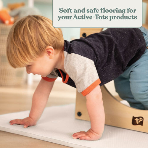 A young child crawls on TP Toys' TP Active-Tots Safety Play Mats beside a wooden play structure, with a sign reading: Soft, non-toxic flooring for your Active-Tots products—ideal for any toddler play area.